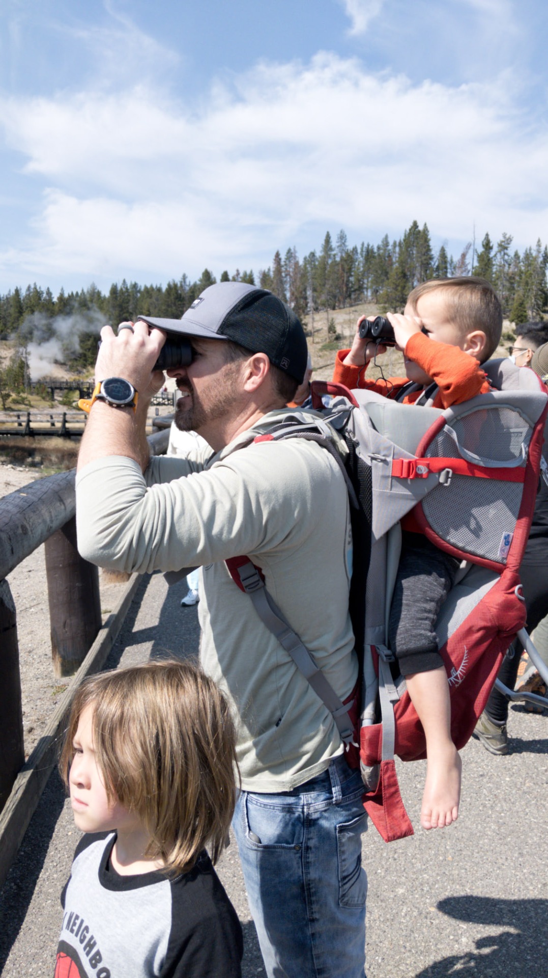 A toddler in a backpack carrier and kids looking through binoculars near a Yellowstone geyser